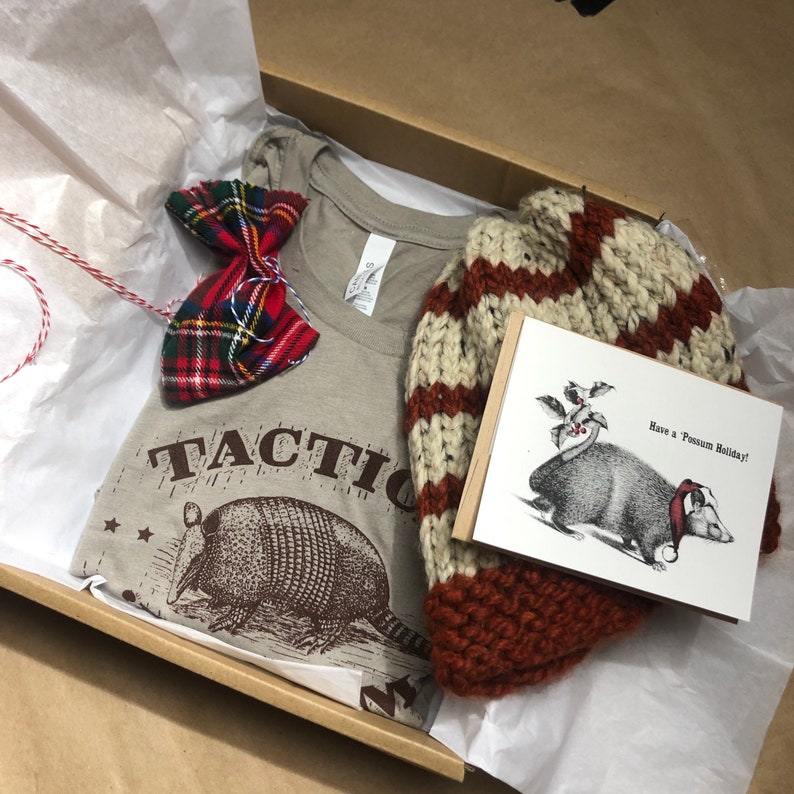 May include: A gift box filled with a brown t-shirt with the text "TACTIC" and a graphic of an armadillo, a red and black plaid bow tie, a brown and white striped knit hat, and a card with the text "Have a Possum Holiday!" and a black and white illustration of a possum.