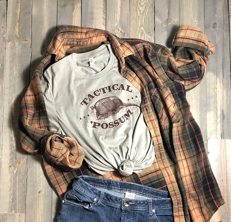 May include: A gray t-shirt with a brown graphic of an armadillo and the text "Tactical Possum". The shirt is layered over a plaid flannel shirt and blue jeans.