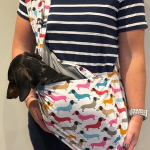 May include: A fabric pet sling carrier featuring a repeating pattern of colourful dachshunds in shades of pink, blue, yellow, and grey on a white background. A small black and tan dachshund is resting inside the pouch of the sling.