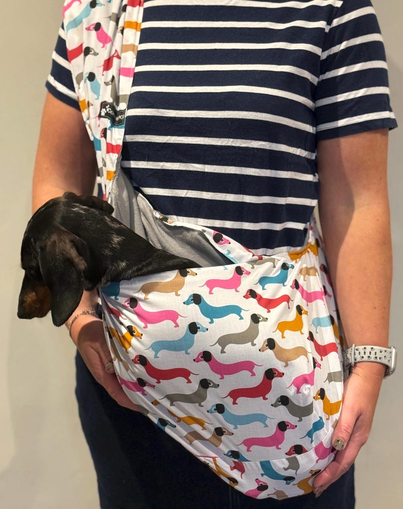 May include: A fabric pet sling carrier featuring a repeating pattern of colourful dachshunds in shades of pink, blue, yellow, and grey on a white background. A small black and tan dachshund is resting inside the pouch of the sling.