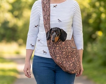 May include: A dark brown fabric pet carrier sling featuring a detailed orange and gold paisley pattern. The sling is worn across the chest and contains a small black and tan dapple dachshund dog with its head visible at the top opening.