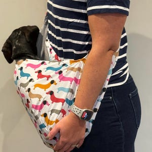 May include: A white fabric pet sling carrier featuring a repeating pattern of colourful dachshund dogs in shades of pink, blue, yellow, and brown. The sling is designed to be worn over the shoulder and contains a small black puppy inside the pouch.