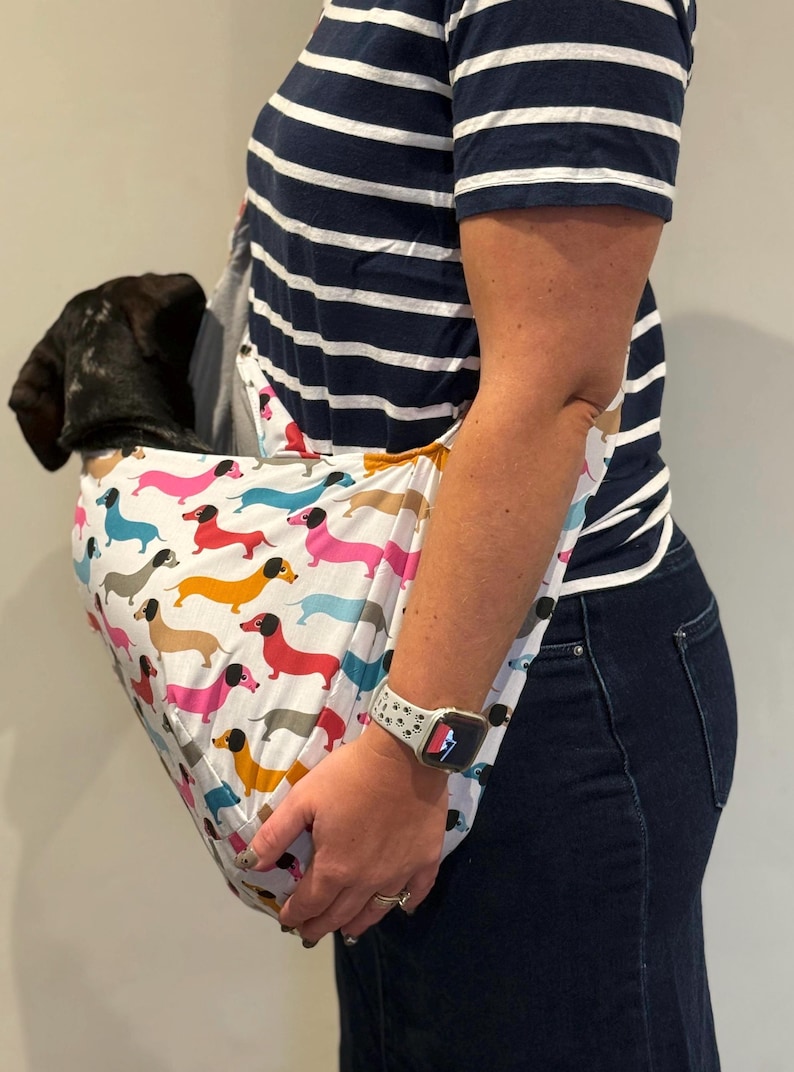 May include: A white fabric pet sling carrier featuring a repeating pattern of colourful dachshund dogs in shades of pink, blue, yellow, and brown. The sling is designed to be worn over the shoulder and contains a small black puppy inside the pouch.