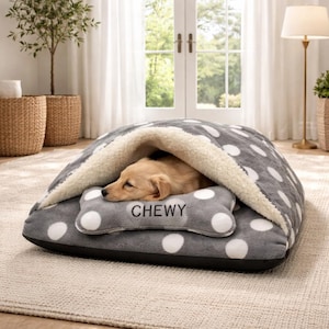 May include: A gray and white polka dot dog bed with a cozy, hooded design. The bed features a bone-shaped pillow with the word "CHEWY" embroidered on it. A small, light brown dog is resting inside the bed.