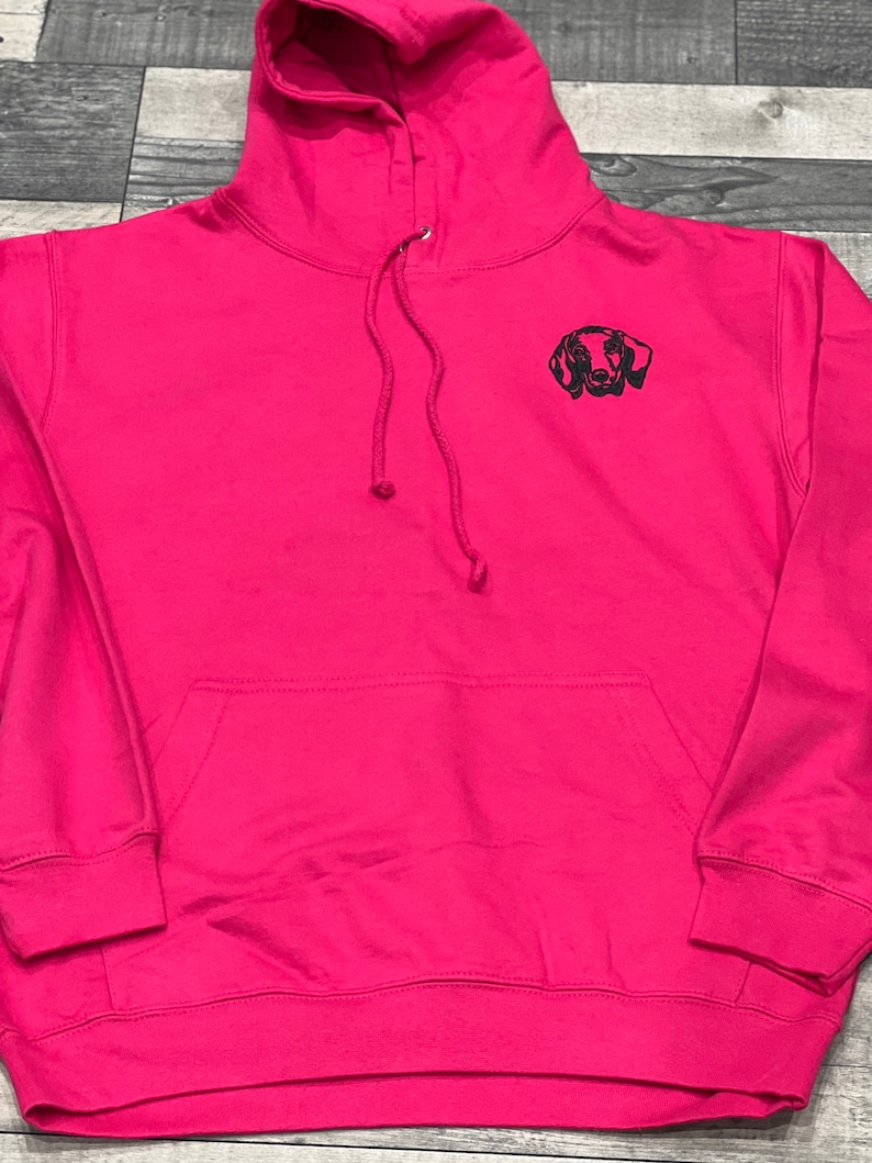 Op de afbeelding: Een roze hoodie met een geborduurd zwart dackel silhouet op de voorkant.