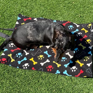 May include: A black and tan dachshund resting on a rectangular black pet mat featuring a colorful pattern of red, yellow, and light blue bones and paw prints. The mat includes black fabric straps attached to one side for carrying or hanging.