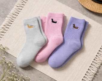 Op de afbeelding: Drie paar enkelsokken in pastelkleuren: grijs, roze en lavendel. Elke sok heeft een geborduurd teckel. De sokken zijn gerangschikt op een crèmekleurige geweven mat.