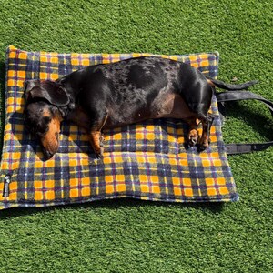 May include: A black and tan dachshund with a dapple coat pattern lying on a rectangular pet mat. The mat features a yellow, navy blue, and white plaid pattern with a soft texture and includes a black fabric carrying strap attached to one side.