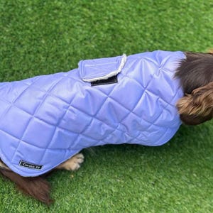 May include: A light purple quilted dog coat with a rectangular pocket and a white trim. The coat is on a dog with brown fur, lying on green grass. The coat has a label that says "Coralia K9".