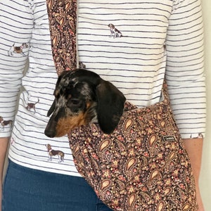 Op de afbeelding: Een bruine en zwarte paisley-gepatroneerde hondendrager met een kleine teckel erin. De drager heeft een lange band en wordt over de schouder gedragen.