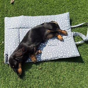 May include: A black and tan dachshund lying on a rectangular, quilted pet mat. The mat features a light gray fabric with a repeating pattern of small white dog silhouettes and includes two attached fabric carrying straps on the right side.