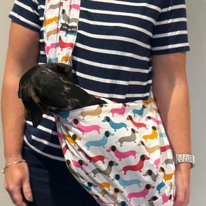 May include: A white fabric pet sling carrier featuring a repeating pattern of colourful dachshunds in shades of pink, blue, yellow, and grey. The sling has a black label with the text Cornish K9 and contains a small black and grey dachshund.