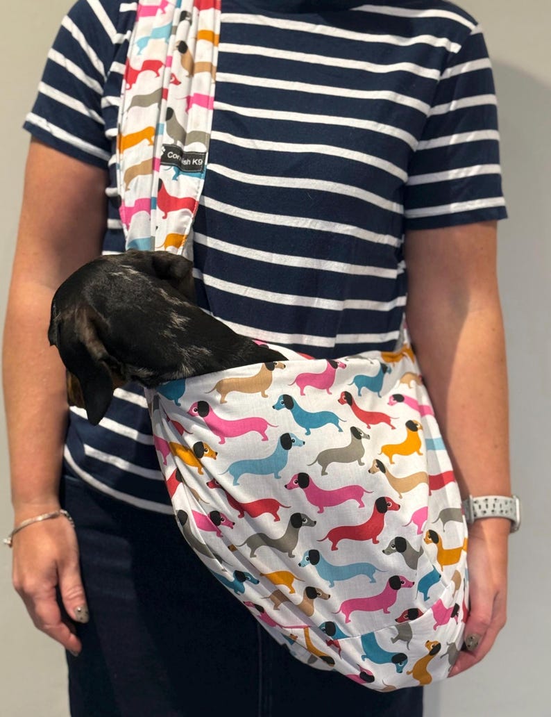 May include: A white fabric pet sling carrier featuring a repeating pattern of colourful dachshunds in shades of pink, blue, yellow, and grey. The sling has a black label with the text Cornish K9 and contains a small black and grey dachshund.