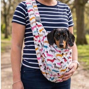 May include: A white fabric pet sling carrier featuring a repeating pattern of colourful dachshund dogs in shades of pink, blue, yellow, and brown. The sling is worn across the chest and contains a black and tan dachshund dog looking forward.