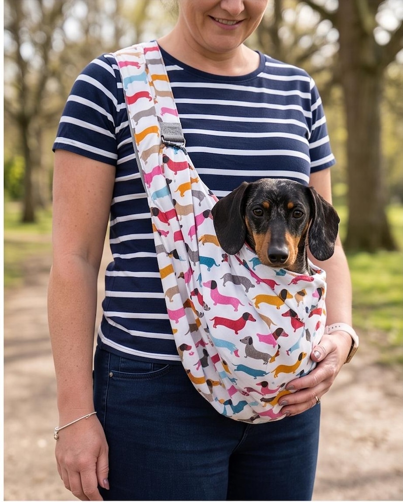May include: A white fabric pet sling carrier featuring a repeating pattern of colourful dachshund dogs in shades of pink, blue, yellow, and brown. The sling is worn across the chest and contains a black and tan dachshund dog looking forward.
