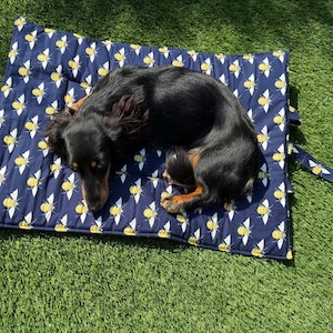 May include: A rectangular quilted pet mat featuring a dark blue fabric with a repeating yellow and white bee pattern. A black and tan long-haired dachshund is curled up in the center of the mat, which includes a matching fabric strap attached.