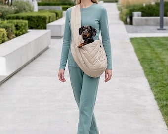 May include: A beige quilted fabric pet carrier sling worn across the torso, containing a small dapple-patterned dachshund dog with a black collar. The carrier features a wide shoulder strap and a soft, padded construction for carrying a small animal.