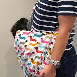 May include: A white fabric pet sling carrier featuring a repeating pattern of colourful dachshund dogs in shades of pink, blue, yellow, and grey. The sling is worn across the body, containing a small dark-coloured dog resting inside the pouch.