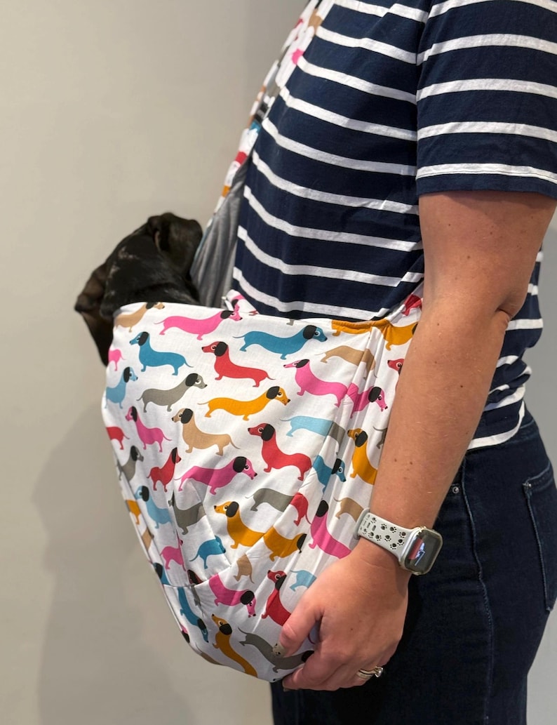 May include: A white fabric pet sling carrier featuring a repeating pattern of colourful dachshund dogs in shades of pink, blue, yellow, and grey. The sling is worn across the body, containing a small dark-coloured dog resting inside the pouch.