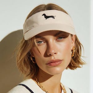 May include: A beige visor with a black dachshund dog silhouette embroidered on the front. The visor has a curved brim and a wide, adjustable band. The visor is made of a soft, lightweight material.