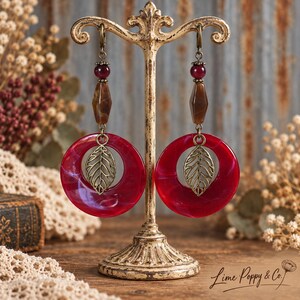 May include: A pair of dangle earrings featuring a large red circular ring with a brass-toned leaf charm suspended in the center. Above the ring is a brown faceted bead and a smaller dark red bead, all attached to a brass-toned hook.