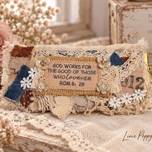 Puede incluir: Un estuche rectangular de tela con un parche central beige que lleva el texto "GOD WORKS FOR THE GOOD OF THOSE WHO LOVE HIM ROM 8: 28". El estuche está decorado con encaje crema superpuesto, retazos de mezclilla, tela marrón y pequeños adornos florales.