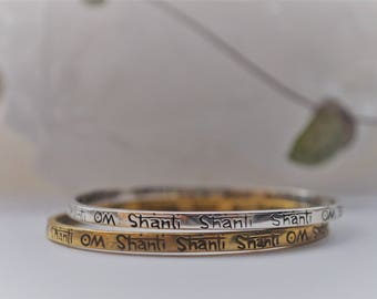 以下が含まれることがあります： 「Om Shanti Shanti Shanti Om」という刻印がそれぞれに繰り返されている、薄い銀と金の2つのバングルです。