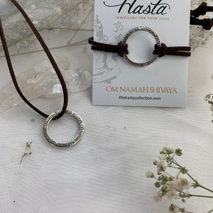 May include: A silver necklace with a circular pendant inscribed with the phrase "Om Namah Shivaya" on a brown leather cord. The necklace is displayed on a white surface with a card that reads "Hasta Jewellery for your soul" and "Om Namah Shivaya" with the website "thehastacollection.com".