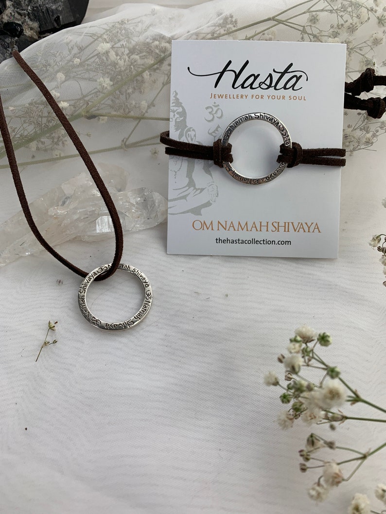 May include: A silver necklace with a circular pendant inscribed with the phrase "Om Namah Shivaya" on a brown leather cord. The necklace is displayed on a white surface with a card that reads "Hasta Jewellery for your soul" and "Om Namah Shivaya" with the website "thehastacollection.com".