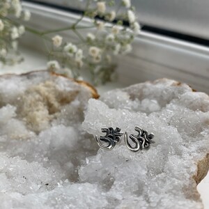 May include: Two silver Om symbol earrings on a white crystal background.