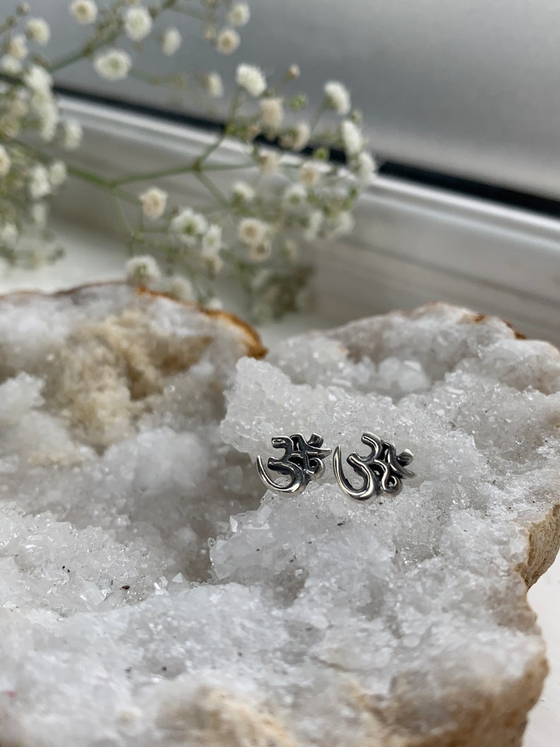 May include: Two silver Om symbol earrings on a white crystal background.