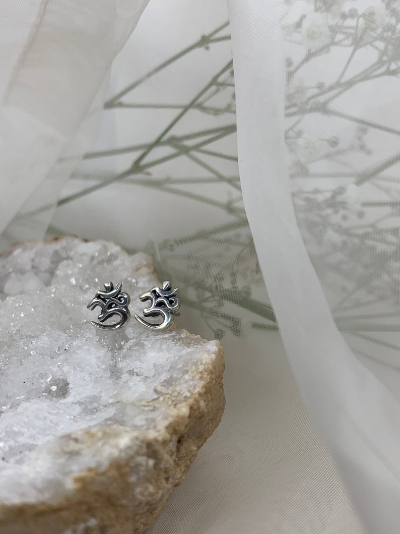 May include: A pair of silver Om symbol stud earrings on a white crystal rock.