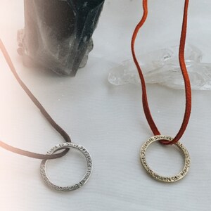 May include: Two silver and gold necklaces with inscribed rings. The silver ring says "Consciouness of the Personification of Shiva". The gold ring says "Namah Shivaya Namah Shivaya". The necklaces are on a white surface with a black crystal and a clear crystal.