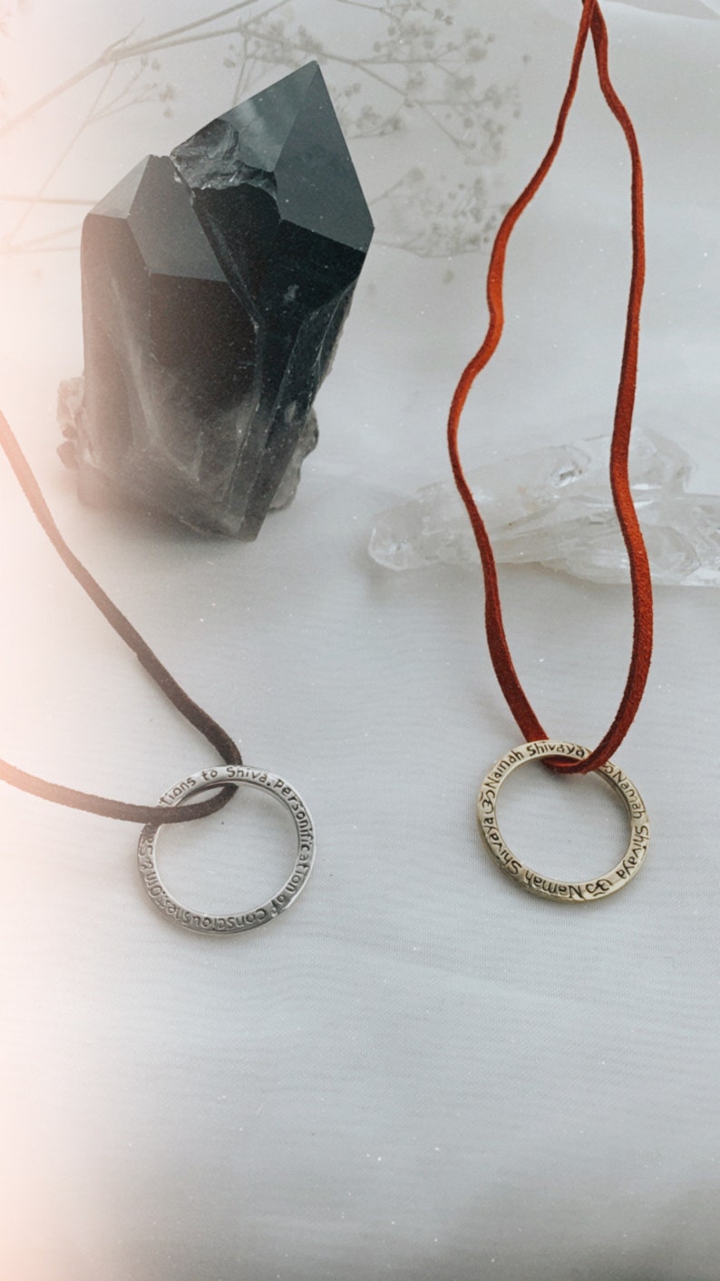 May include: Two silver and gold necklaces with inscribed rings. The silver ring says "Consciouness of the Personification of Shiva". The gold ring says "Namah Shivaya Namah Shivaya". The necklaces are on a white surface with a black crystal and a clear crystal.
