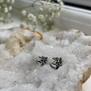 May include: A pair of silver Om symbol earrings on a white crystal background.