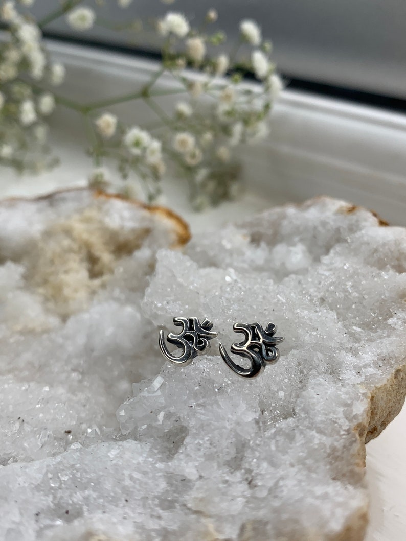 May include: A pair of silver Om symbol earrings on a white crystal background.