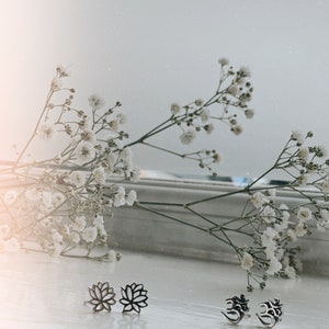 May include: A set of silver earrings featuring a lotus flower design and the Om symbol. The earrings are displayed on a white surface with white flowers in the background.