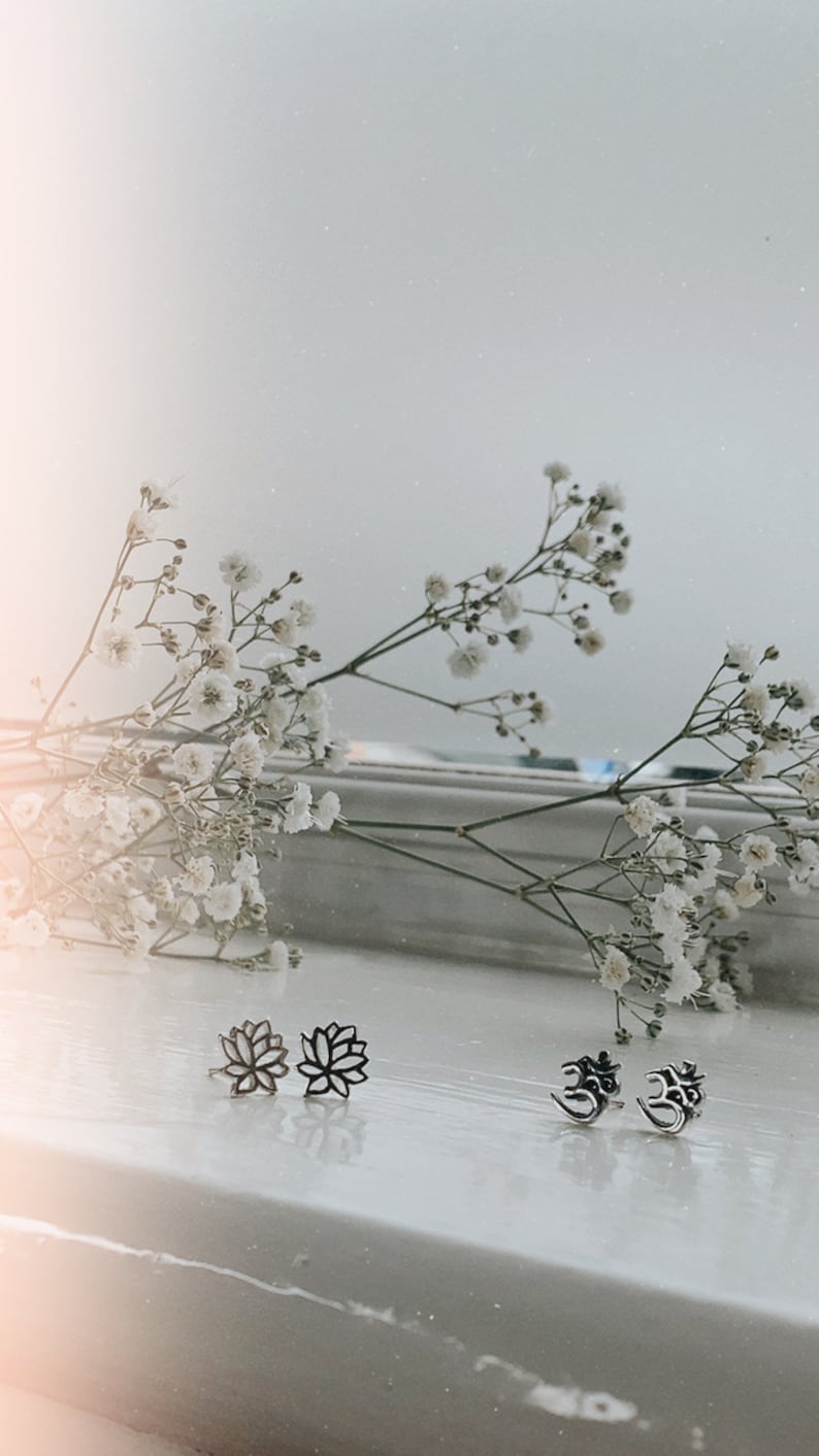 May include: A set of silver earrings featuring a lotus flower design and the Om symbol. The earrings are displayed on a white surface with white flowers in the background.