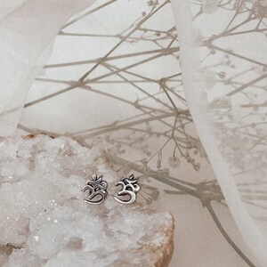 May include: A pair of silver Om symbol earrings on a white crystal rock with a sheer white fabric background.