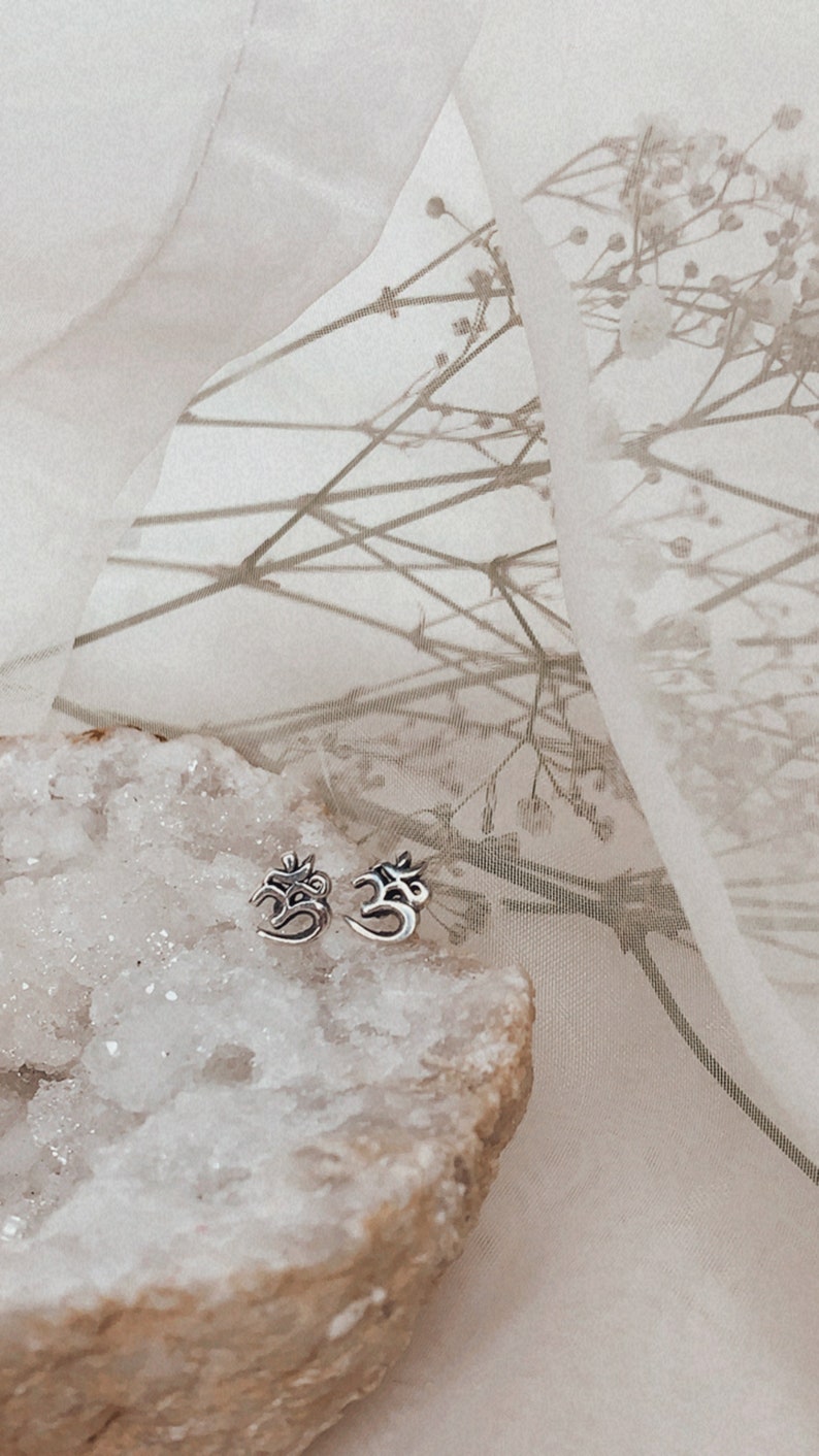 May include: A pair of silver Om symbol earrings on a white crystal rock with a sheer white fabric background.