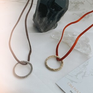 May include: Two necklaces with inscribed circular pendants on a white surface. The necklaces are on brown and red cords. A black crystal and a clear crystal are in the background. The text on the card reads "Om Namah Shivaya" and "thehastacollection.com".