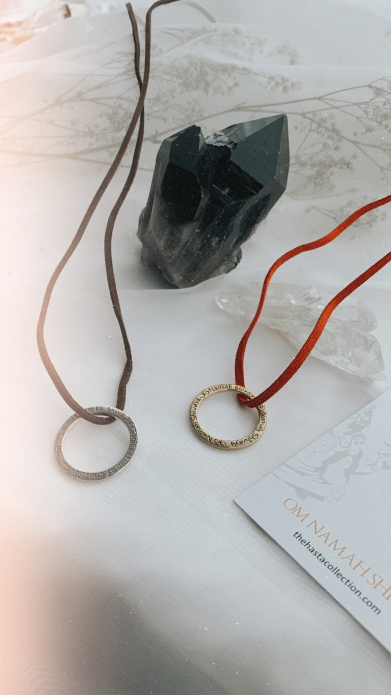 May include: Two necklaces with inscribed circular pendants on a white surface. The necklaces are on brown and red cords. A black crystal and a clear crystal are in the background. The text on the card reads "Om Namah Shivaya" and "thehastacollection.com".