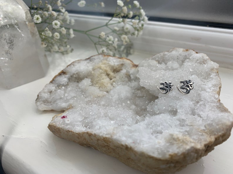 May include: A pair of silver Om symbol earrings displayed on a white crystal geode.