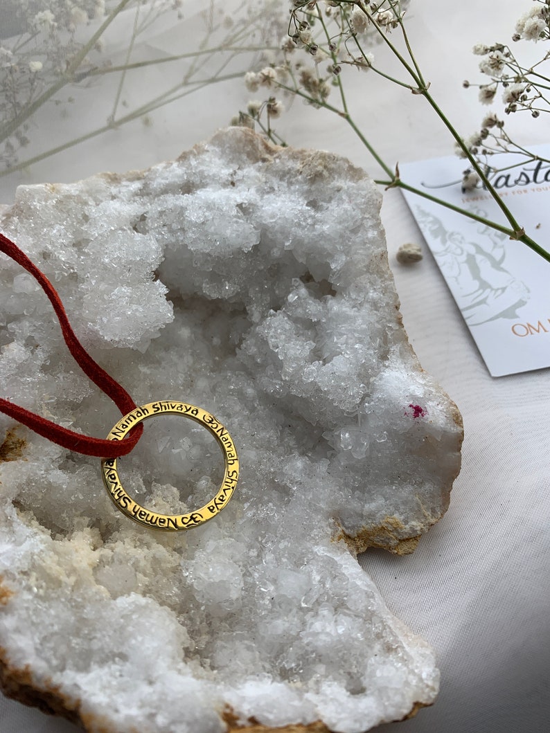 May include: A gold-toned necklace with a circular pendant inscribed with the phrase "Namah Shivaya" in Sanskrit. The necklace is displayed on a white crystal rock.
