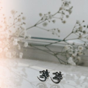 May include: A pair of silver earrings with an Om symbol design. The earrings are on a white surface with a blurred background of white flowers.