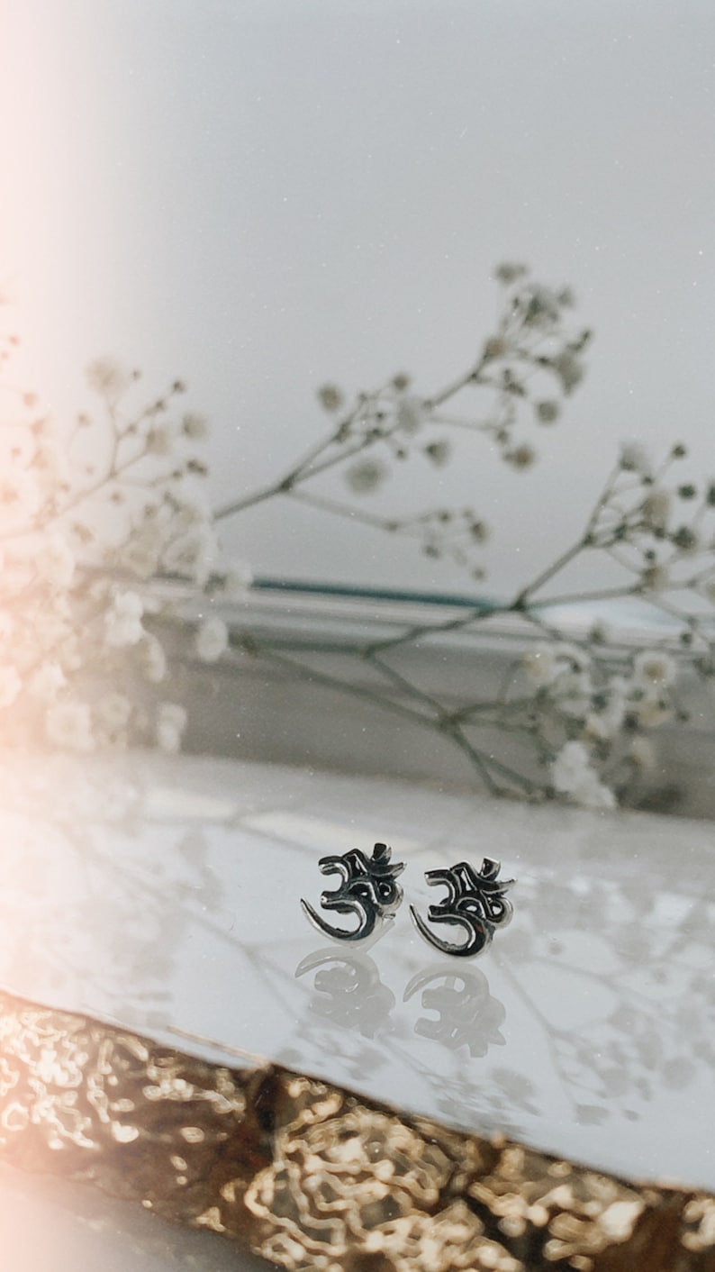 May include: A pair of silver earrings with an Om symbol design. The earrings are on a white surface with a blurred background of white flowers.