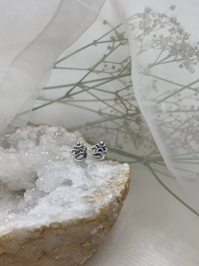 May include: A pair of silver Om symbol earrings on a white crystal rock. The earrings are small and delicate, and the Om symbol is clearly visible.