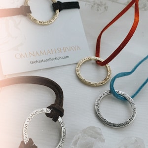 May include: Three silver and gold necklaces with the phrase "Om Namah Shivaya" inscribed on them. The necklaces are on a white surface with a crystal next to them. The necklaces are on different colored cords: black, red, and blue.