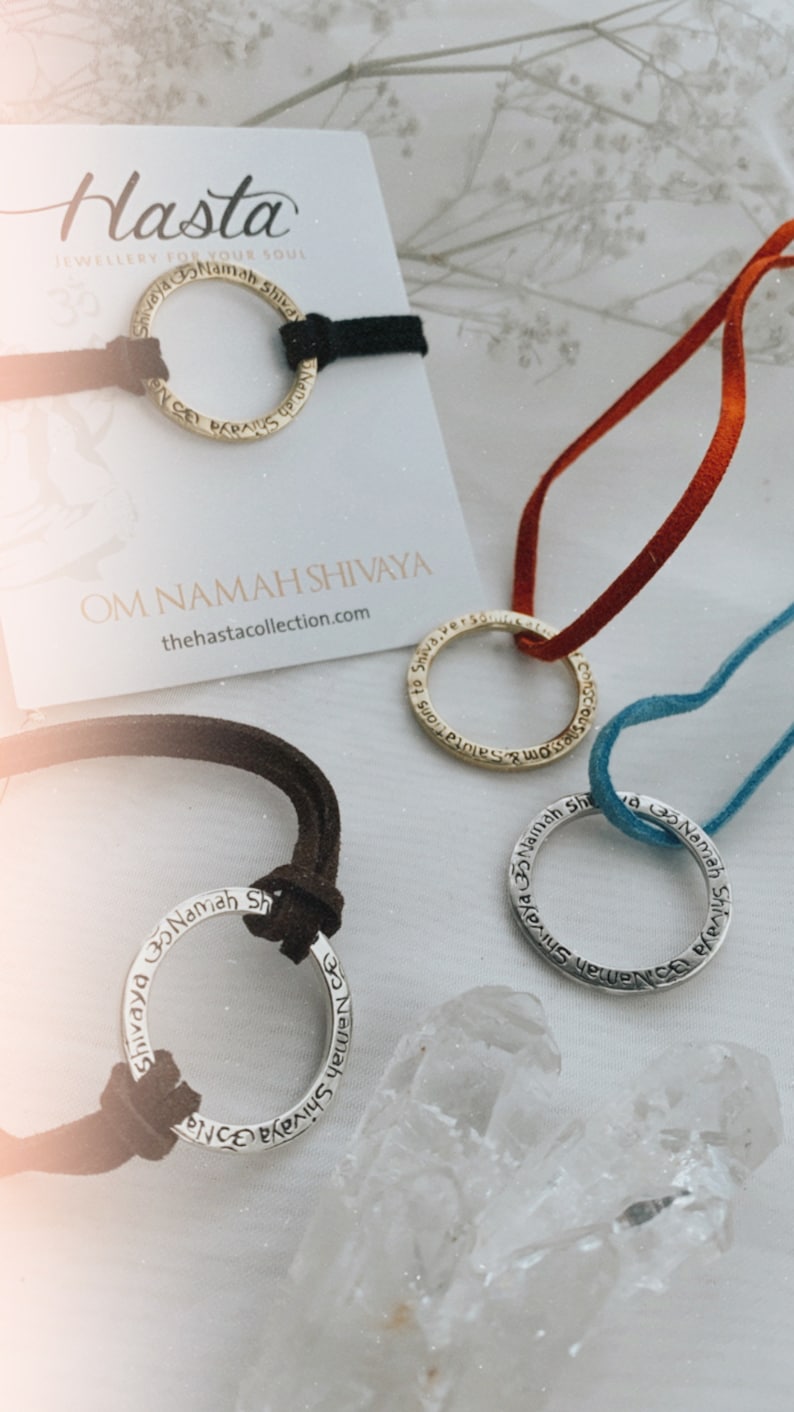 May include: Three silver and gold necklaces with the phrase "Om Namah Shivaya" inscribed on them. The necklaces are on a white surface with a crystal next to them. The necklaces are on different colored cords: black, red, and blue.