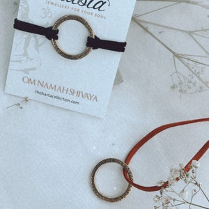 May include: A black cord bracelet with a gold-toned circular charm inscribed with the phrase "Om Namah Shivaya". The bracelet is displayed on a white card with the brand name "Hasta" and the tagline "Jewellery for your soul".